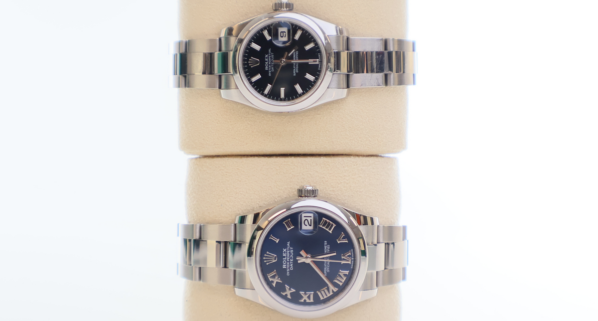 Two Rolex wristwatches with stainless steel bracelets and black and blue dials displayed on a beige stand